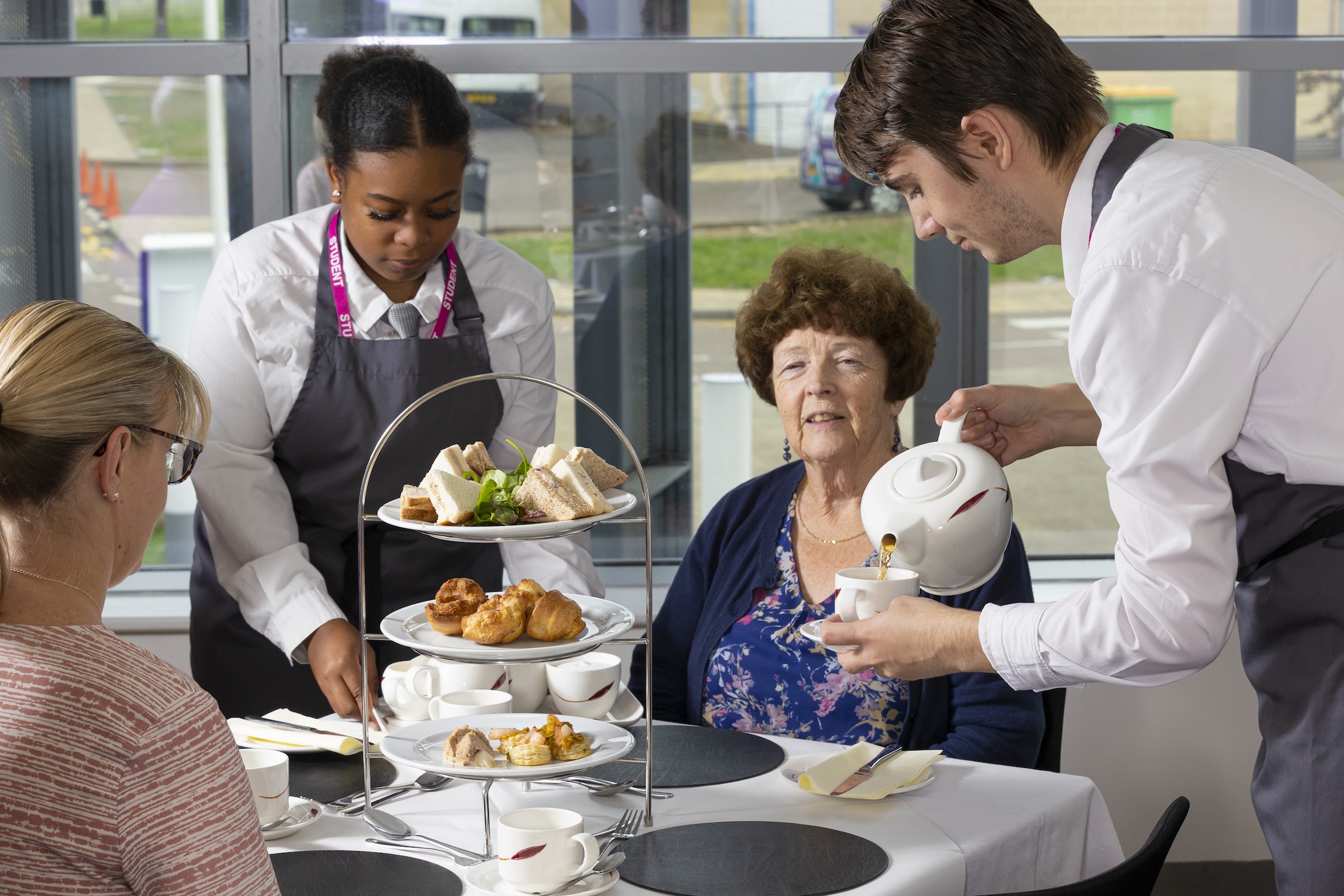 Two students serve tea to two people seated at table